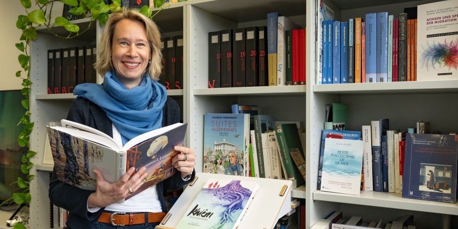 Eine Frau steht vor einem großen Bücherregal und blättert durch ein Buch, welches Sie in ihren Händen hält