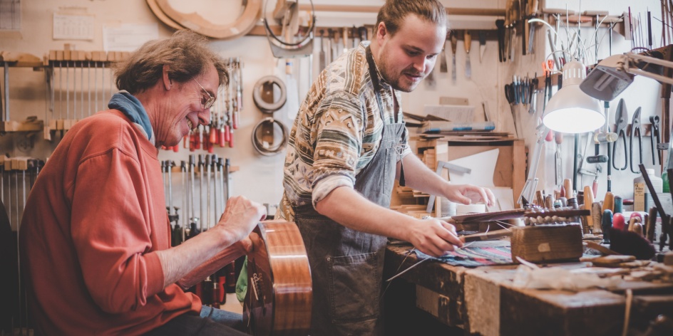 Ein älterer und jüngerer Mann arbeiten gemeinsam an der Werkbank an einer Gitarre.