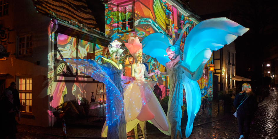 Costumed stilt walkers stand in front of the colourfully illuminated house facades in the Schnoor.