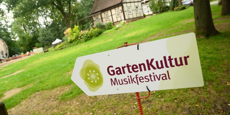 Ein Schild mit der Aufschrift "Gartenkultur Musikfestival" weist den Weg in den Garten. Im Hintergrund befinden sich Bauernhäuser.