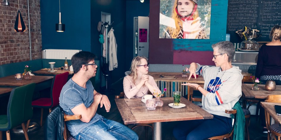 Zwei Frauen und ein Mann sitzen an einem Tisch in einem Café. Eine der Frauen spricht in Gebärdensprache.