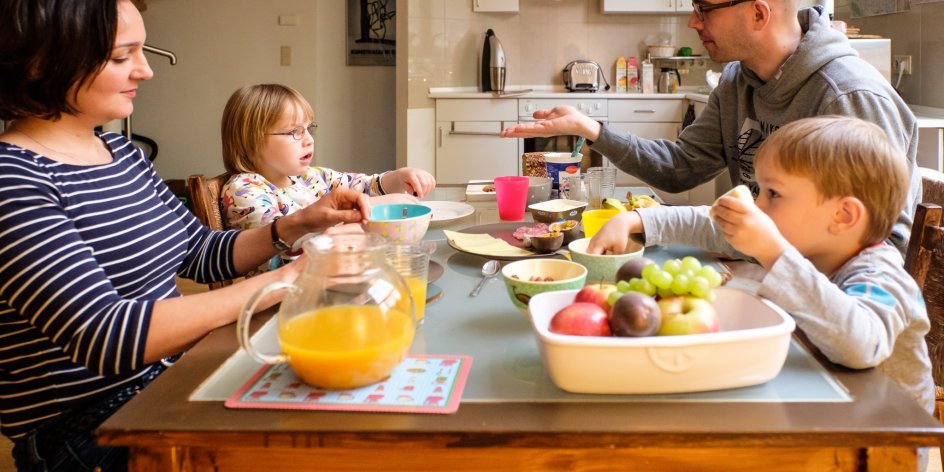 Ein Mann und eine Frau sitzen mit zwei kleinen Kindern am Frühstückstisch. Ein Bild über den Alltag in einer Familie mit behinderten und nicht behinderten Kindern.