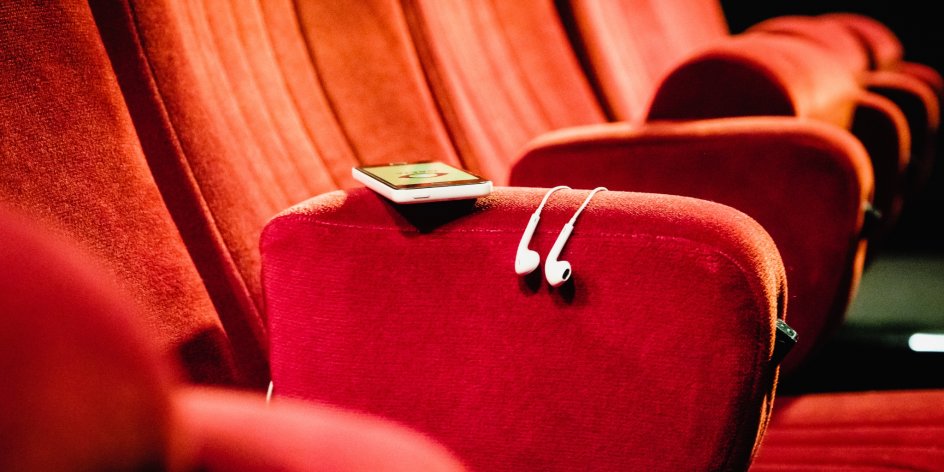 Rote Kinositze, auf einem Sitz liegt ein Smartphone mit Kopfhörern. Auf dem Smartphone ist die GRETA App geladen.