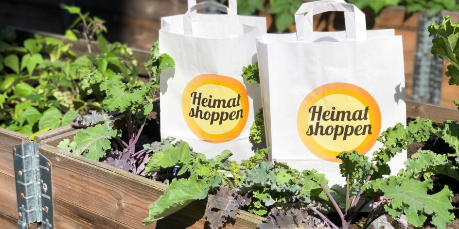 Man sieht zwei Papiertüten mit dem Logo der Aktion "Heimat Shoppen", die in einem Beet stehen.
