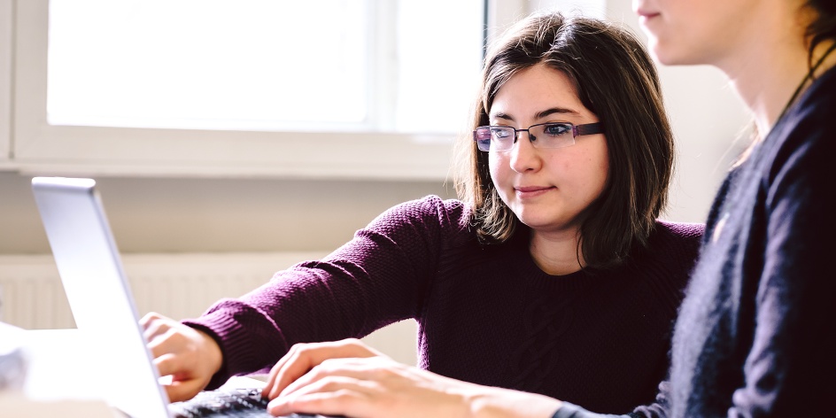 Zwei Frauen sitzen an einem Laptop. Eine tippt auf der Tastatur, die andere zeigt auf den Bildschirm.