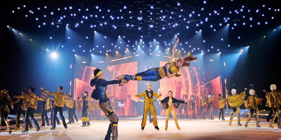 Ein Eiskunstläufer wirbelt eine andere Eiskunstläuferin umher, alle haben glitzernde Kostüme an. 