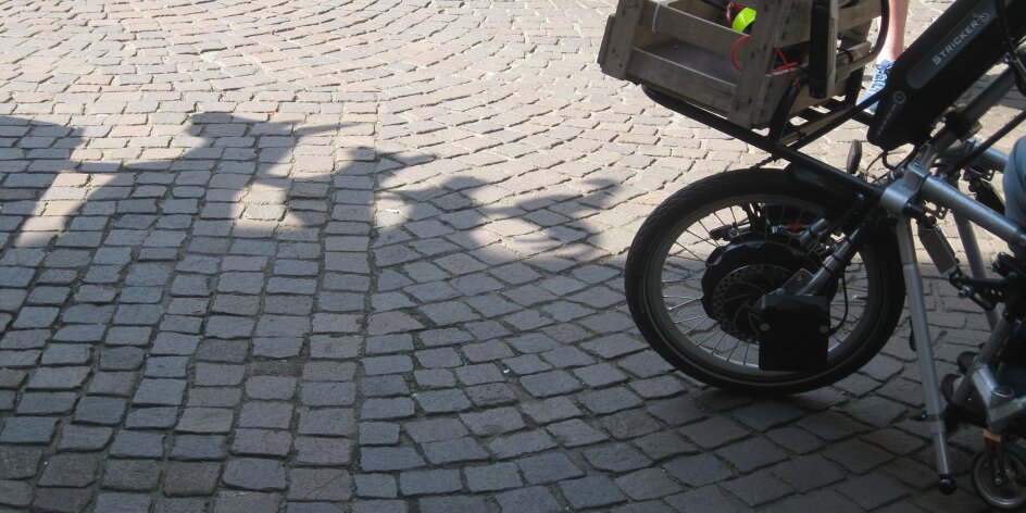 Vorderreifen eines Handbikes. Der Boden ist mit Kopfsteinpflaster versehen, man sieht einen Schatten der Bremer Stadtmusikanten.