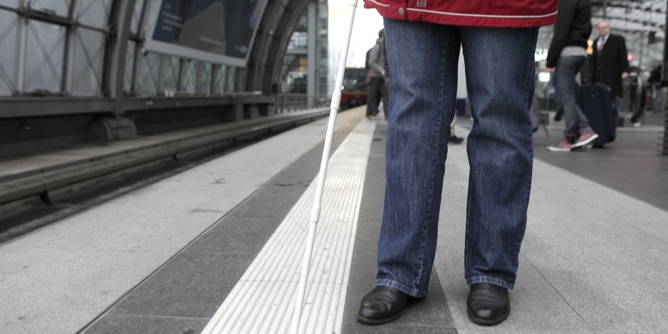Eine Person steht auf einem Bahnsteig und ertastet mit einem Langstock das Bodenleitsystem. 