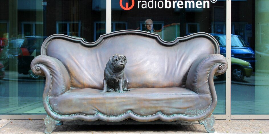 Auf einer Couch aus Bronze sitzt eine Mopsstatue.