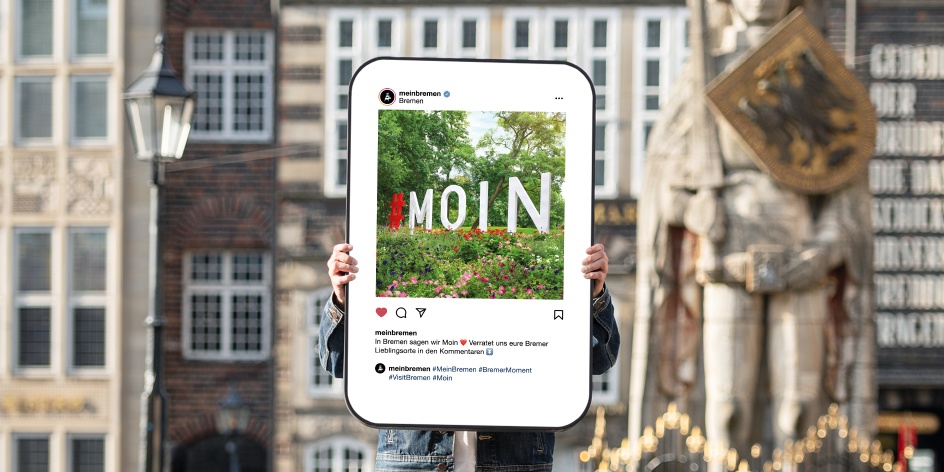 In der Mitte des Bildes wird eine Art Smartphone-Bildschirm von zwei Händen hochgehalten - darauf zu seine ist ein Social Media Post, der ein Foto eines MOIN-Schriftzuges auf einer Blumenwiese zeigt. Hierbei handelt es sich um ein Exponat einer Ausstellung. Im Hintergrund ist unscharf der Bremer Marktplatz mit Roland zu sehen.