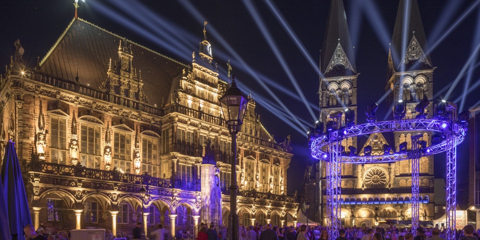 Der beleuchtete Bremer Dom und das beleuchtete Bremer Rathaus bei Nacht. Davor strahlen zahlreiche Scheinwerfer in den Himmel.