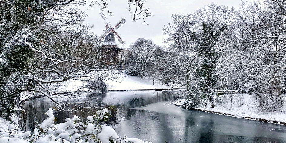 Eine eingeschneite Winterlandschaft ("Wallanlagen") mit einem zugefrorenem Wasserlauf. Im Hintergrund steht die eingeschneite "Mühle am Wall".