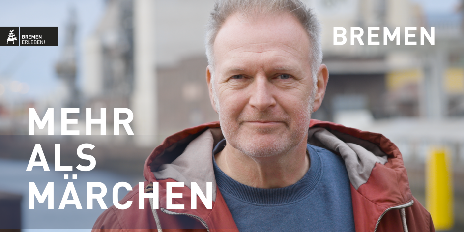 Ein Mann mit roter Jacke und blauem Shirt lächelt in die Kamera. Im Hintergrund ist unscharf der Holz- und Fabrikenhafen in Bremen zu sehen. In der linken, unteren Bildecke steht in weißer Schrift: "MEHR ALS MÄRCHEN". In der rechten, oberen Bildecke steht in weißer Schrift: "BREMEN".