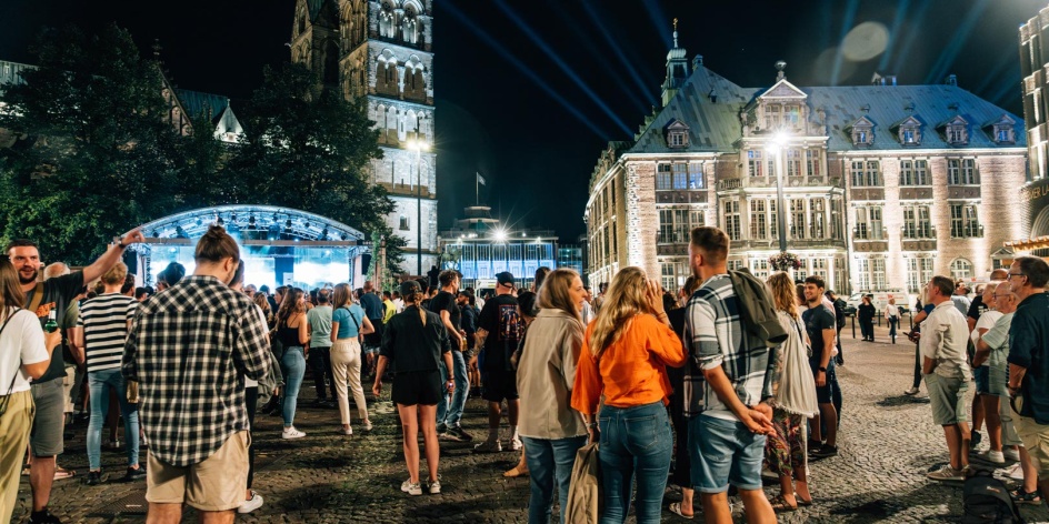Menschen stehen in Grüppchen bei Nacht auf dem Domshof in Bremen vor einer großen Bühne.
