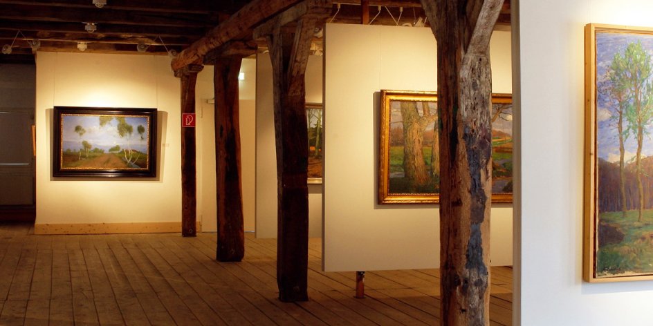 Ein Ausstellungssaal mit Gemälden, Dielenboden und Holzbalken.