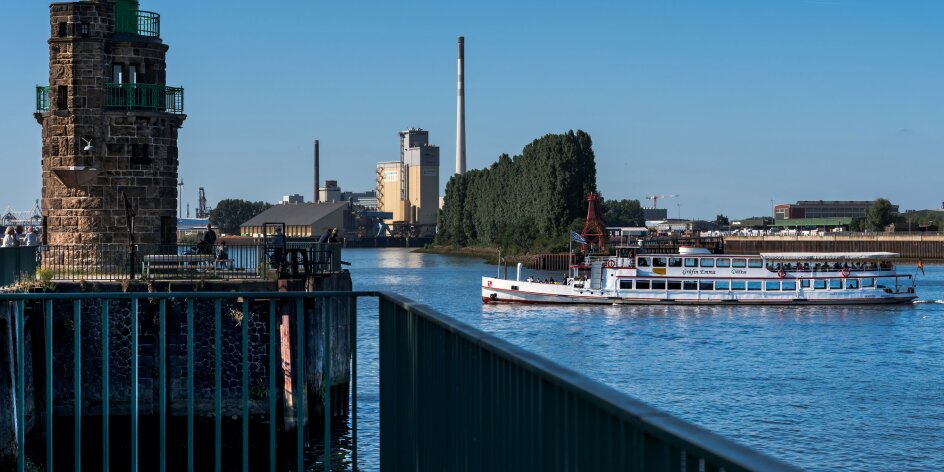 Der Molenturm ragt links im Bild in den blauen Himmel und im rechten Abschnitt fährt auf der Weser ein Schiff.