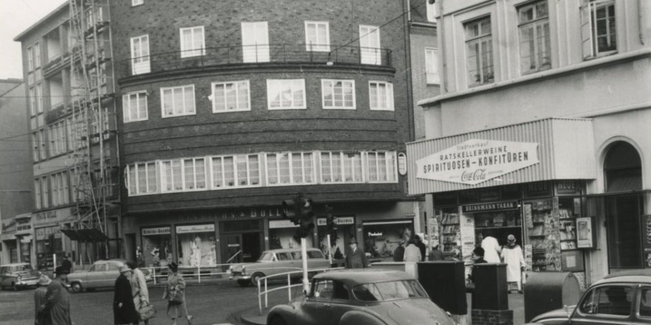 Eine Schwarz-Weiß-Aufnahme einer belebten Straßenkreuzung, der "Sielwallkreuzung", welche von Gebäuden gesäumt ist. Menschen und Autos passieren die Kreuzung.