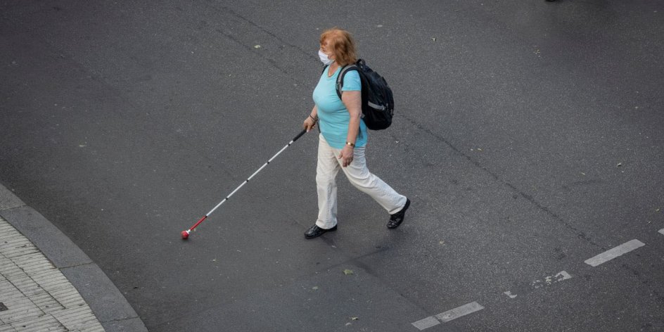 Eine Frau mit Sehbeeinträchtigung kreuzt eine Straße und orientiert sich mit einem Langstock. Sie trägt einen Mund-Nase-Schutz.