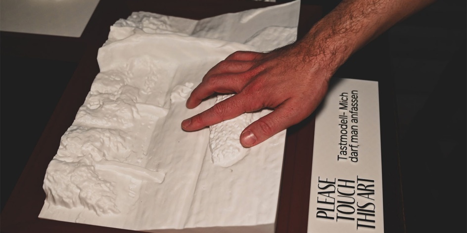 Eine Hand ertastet ein Tastmodell. Darunter ist ein Schild mit der Aufschrift: "Please touch this art. Tastmodell - mich darf man anfassen."