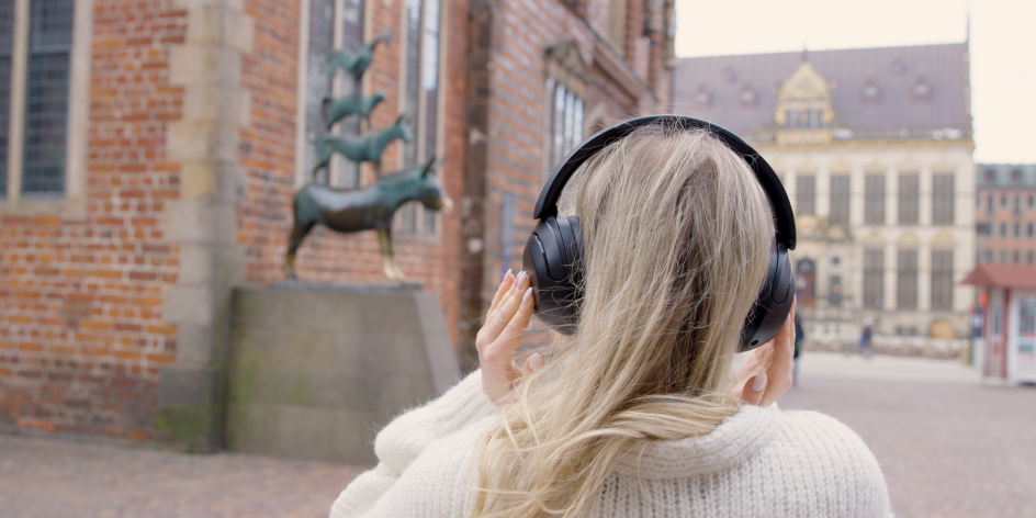 Eine Person mit Kopfhörern steht vor den Bremer Stadtmusikanten am Rathaus und hört einen Audiowalk.