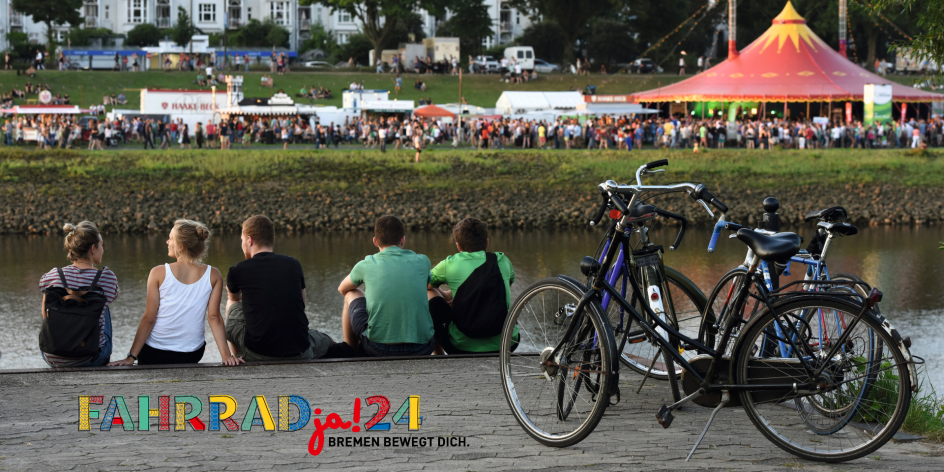 Eine Gruppe sitzt neben Fahrrädern am Ufer. Links ist das Logo von "FAHRRADja! Bremen bewegt Dich." zu sehen. Auf der anderen Flussseite ist ein Festival verschwommen im Hintergrund.