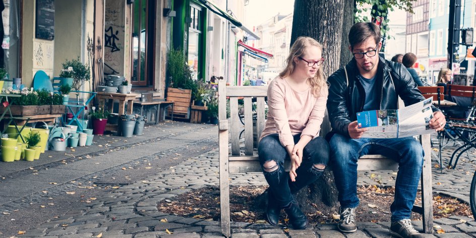 Eine Junge Frau und ein junger Mann sitzen auf einer Holzbank an einer Straße und schauen zusammen auf einen Flyer.