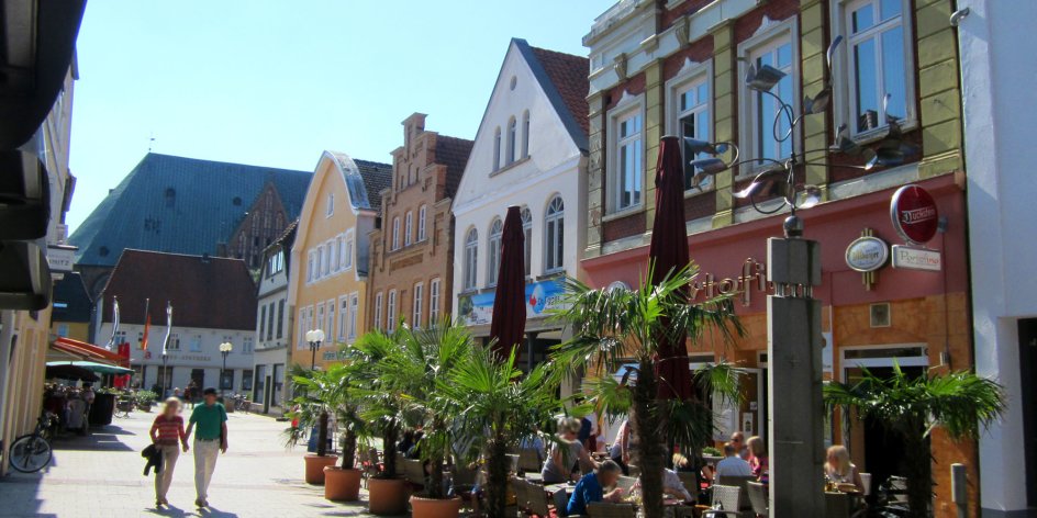 Eine Fußgängerzone mit alten Häusern an einem sonnigen Tag. Vor einem Haus befindet sich ein Straßencafé, umsäumt von Palmen. 