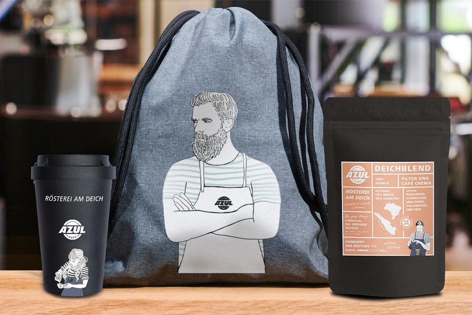 Auf einem Tisch stehen ein schwarzer Mehrweg-Becher für unterwegs, ein grauer Beutel sowie eine Packung Kaffee der Sorte Deichblend. Alle Produkte sind von Azul Rösterei am Deich.