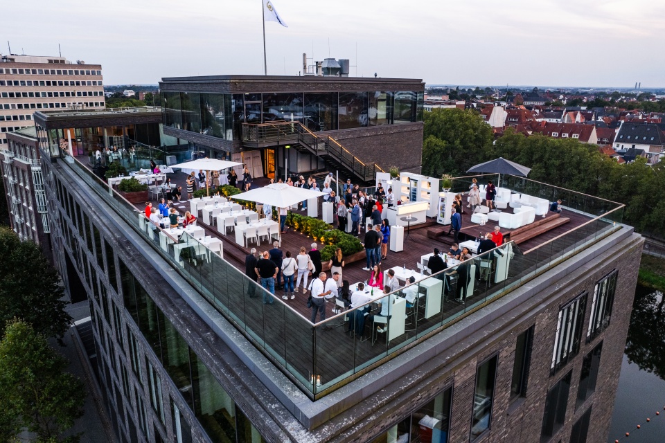 Luftaufnahme von einem großen Gebäude auf dem Teerhof mit Blick auf die Dachterrasse, auf der ein Event stattfindet.