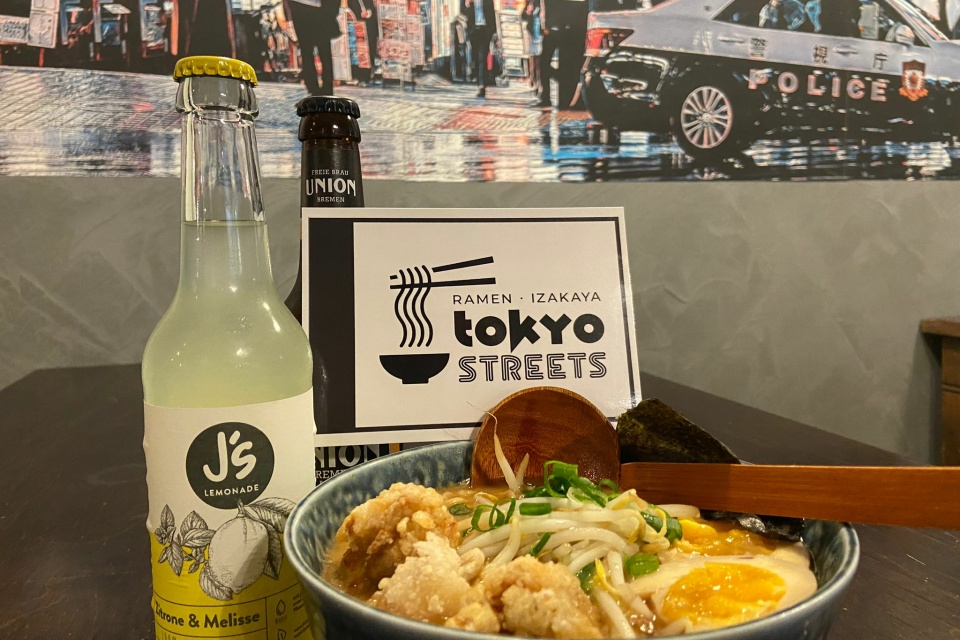 Ein gedeckter Tisch mit einer Schüssel Ramen, einer Limonade, einem Bier und einer Gutscheinkarte von Tokyo Streets