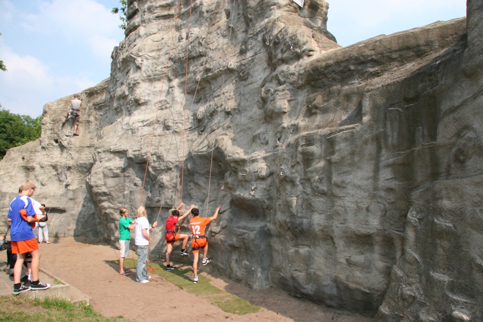 Eine Gruppe von Menschen steht vor einer großen künstlichen Kletterwand im Freien. Zwei Kinder in orangenen Sporttrikots und Klettergurten beginnen gerade, die Wand zu erklimmen, während Erwachsene am Boden die Sicherungsseile halten. Im Hintergrund klettert eine Person höher an der Wand, gesichert durch ein Seil. Die Wand ist grau und hat viele Griffe und Haken für das Klettern. Einige Zuschauer, darunter ein Junge in einem blau-weißen Trikot, beobachten das Geschehen. Der Himmel ist klar mit ein paar Wolken, und es ist sonnig.