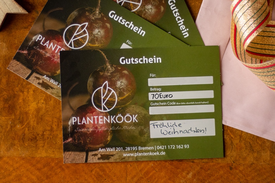 Auf einem Holztisch liegen drei grüne Gutscheine des Restaurants Plantenkök in Höhe von 70 Euro mit einem weihnachtlichen Gruß