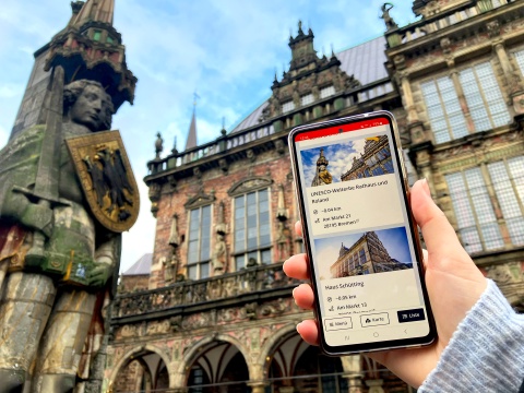 Auf einem Smartphone sieht man Infos zu Rathaus und Roland, die man auch im Hintergrund sieht.