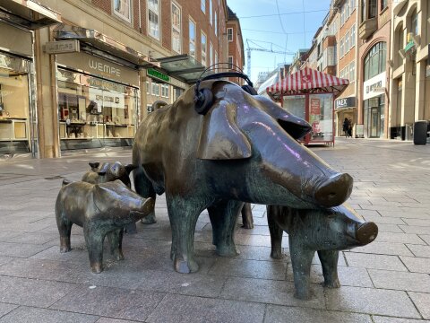 Schweine aus Messing stehen in einer Einkaufstraße. Dem größten Schwein wurden Kopfhörer aufgesetzt. 