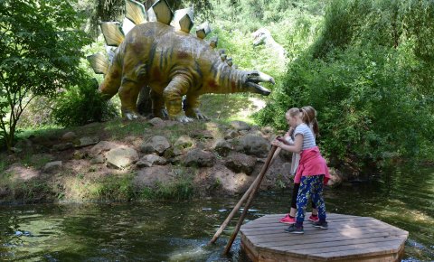 Zwei Mädchen führen ein hölzernes Floß, im Hintergrund das Modell eines Dinosauriers am Ufer.
