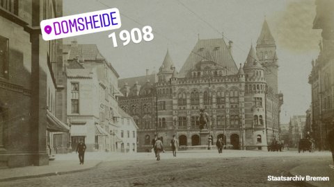 Zu sehen ist ein kleiner Platz mit einer Statue. Am Ende des Platzes steht ein großes Gebäude mit mehreren kleinen Türmen. Links im Bild ist eine angeschnittene Häuserreihe zu sehen. Ein lilafarbener Schriftzug mit der Aufschrift "Domsheide" sowie die weiße Zahl "1908" sind auf das braune Bild gelegt.