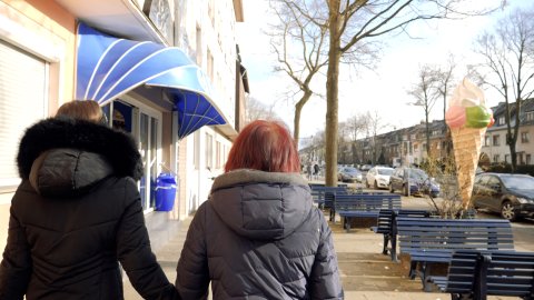 Zu sehen sind zwei weibliche Personen von hinten, die beide eine Winterjacke tragen und an einer Straße entlanggehen. Rechts stehen blaue Bänke und eine große Eiswaffel-Figur. Links ist eine blaue Markise zu sehen.