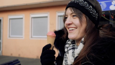 Zu sehen ist eine weibliche Person, die eine Eiswaffel in der Hand hält und lächelt. Die Person trägt ein schwarzes Stirnband, einen weißen schal sowie schwarze Handschuhe.