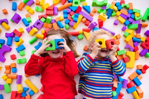 Zwei Kinder halten sich Spielholzklötze vor die Augen und gucken in die Kamera. Um sie herum liegen viele bunte Bauklötze