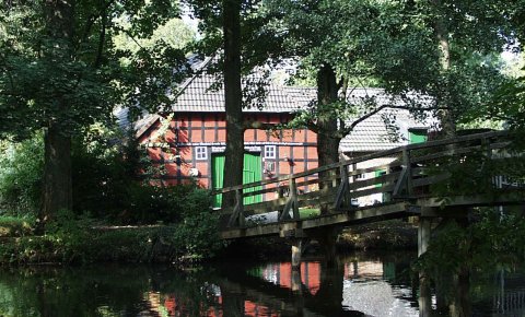 Eine hölzerne Brücke führt über einen Fluss zu einem Fachwerkhaus.