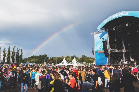 Das MS Dockville Festival mit der Hauptbühne. Davor sind viele Besucher. Am Himmel ist ein Regenbogen.