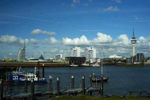 Bremerhavens Skyline ist in Form von Gebäuden und einem Turm im Hintergrund zu sehen. Davor ist ein Wasserkanal zu sehen. Im Vordergrund führt ein Steg zum Wasser. Einige Schiffe sind links im Bild festgemacht.