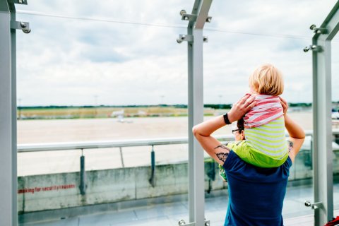 Eine Frau trägt ein Kind auf ihren Schultern. Beide gucken durch eine Scheibe nach draußen auf das Flughafengelände.