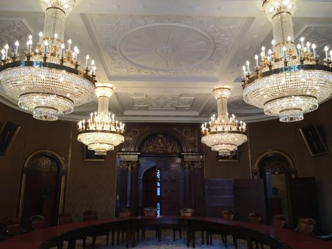 An der Decke hängen vier große Kronleuchter, die mit Diamanten besetzt sind. Die Decke selbst ist weiß und mit Ornamenten verziert. Der Raum wird von einem runden Tisch ausgefüllt, an dem zahlreiche Stühle stehen. Auf der gegenüberliegenden Seite befindet sich eine große hölzerne Tür, die mit goldenen Ornamenten verziert ist.