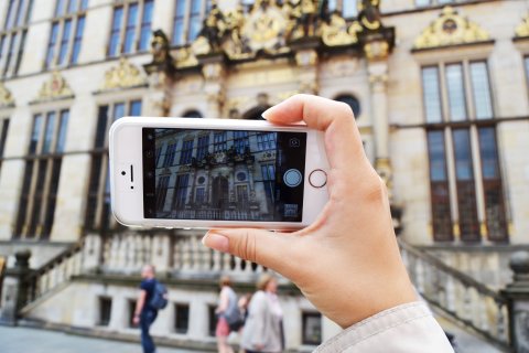 Zu sehen ist eine Hand, die ein Handy hält. Auf dem Handybildschirm ist der Ausschnitt eines Gebäudes zu erkennen. Dieses Gebäude sieht man auch im Hintergrund des gesamten Bildes. Es hat eine goldene Fassade und Treppen.
