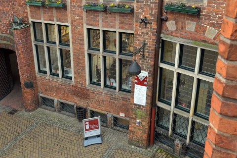 Der Blick von oben fällt auf die Wand eines Backsteingebäudes mit Fenstern. Hinter den Fenstern sind Büroräume zu erkennen. Links ist ein Durchgang in der Backsteinwand zu sehen. 