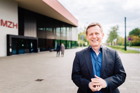 Auf dem Bild ist ein lächelnder Mann zu sehen. Im Hintergrund befindet sich ein Gebäude der Universität Bremen.