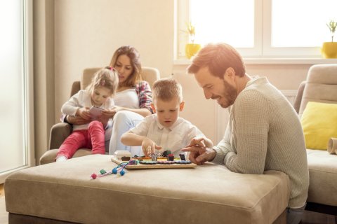 Junge Familie sitzt auf dem Sofa und spielt gemeinsam ein Brettspiel im sonnendurchfluteten Wohnzimmer.