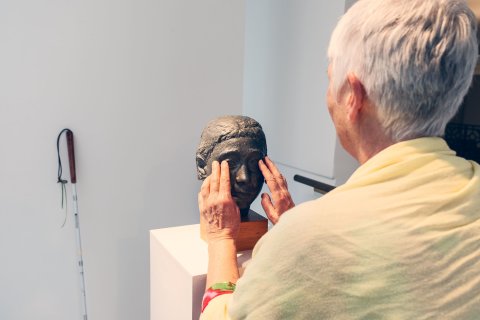 Eine Frau ertastet die Augen einer Bronzefigur in Form eines Frauenkopfes in einer Austellung.