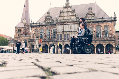 Ein Mann und eine Frau auf dem Marktplatz vor dem Bremer Rathaus. Die Frau sitzt im Rollstuhl.